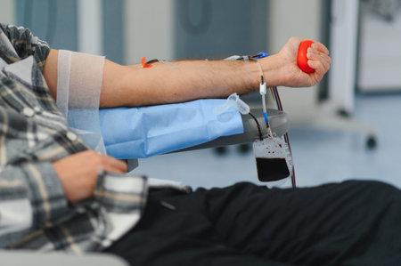 Blood donor sitting, arm extended, and receiving a needle for blood collection into bag, holding a...