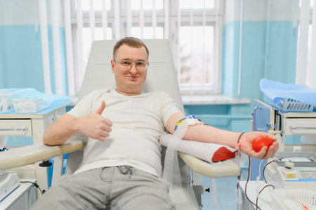 Man donating blood in a modern clinic, showing arm with needle and holding a stress ball, confidently giving a thumbs up gestureの写真素材