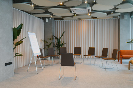 Empty modern meeting room featuring a circle of chairs, flipchart, and acoustic ceiling panels,...