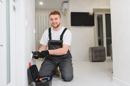 Professional electrician wearing overalls and gloves, installing a new electrical outlet with tools...