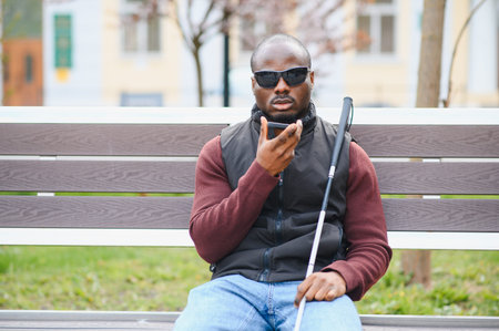 Blind african american man with a white cane sitting on a park bench, using his smartphone's voice assistant