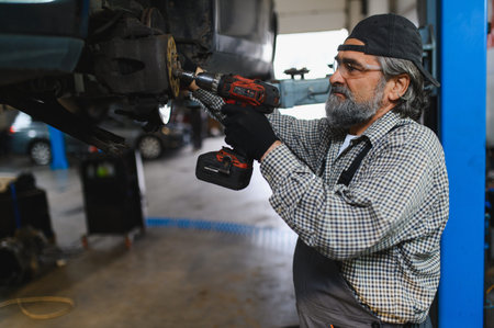 Experienced auto mechanic in overalls using an electric drill to service a vehicle's disc brake in a professional garage setting