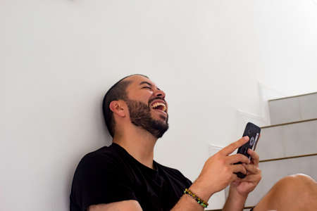 Latin man sitting on the stairs, laughing loudly with phone in his hands. Smiling man. Concept of...