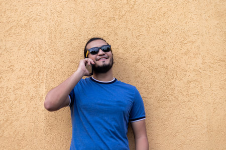 young latino man with sunglasses in a phone call. Standing at a yellow wall. Technology concept