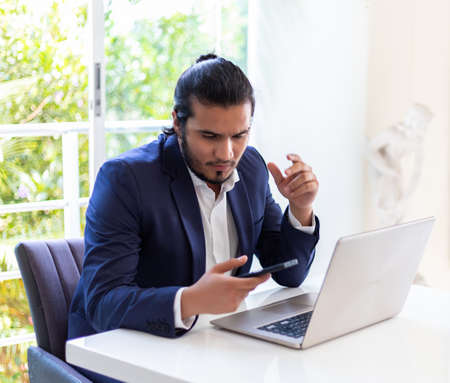 young latino Man with laptop texting on mobile phone sitting at table. Home workingの写真素材