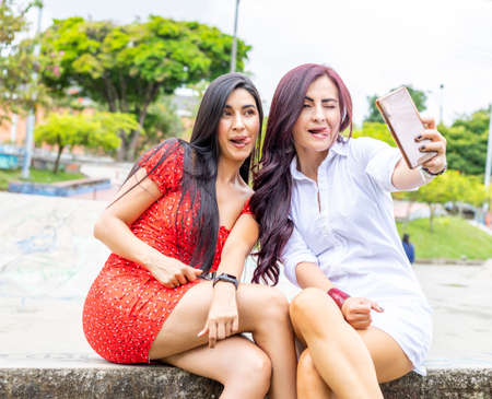 Two Smiling cute young girl friends outdoors on public bench take a selfie with smart phone. Shows tongueの写真素材