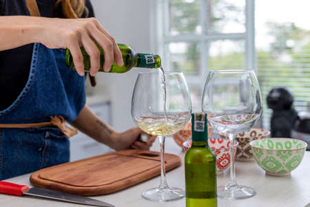 woman in the kitchen pouring two glasses of white wine while she cooksの写真素材