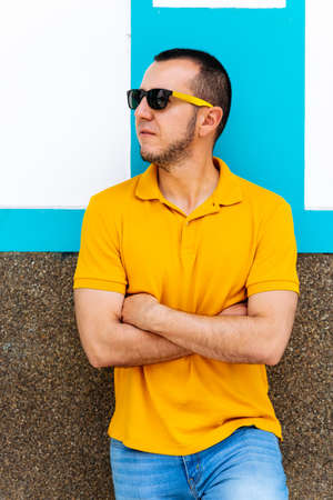 vertical shot of a young latino man with sunglasses and crossed arms against a blue and white colored wall.の写真素材