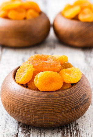 Dried apricots in wooden bowls on a rustic white wooden table, the bright orange color of the apricots contrasting with the natural brown of the bowls.の写真素材