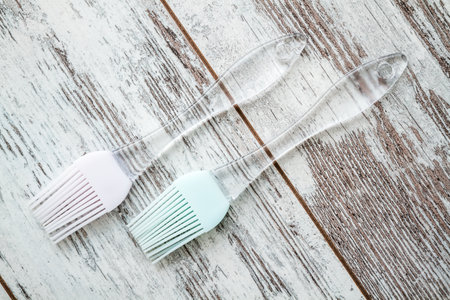 Two silicone basting brushes with transparent handles, one with a pink head and the other with a light blue head, placed on a rustic wooden white surface.の写真素材