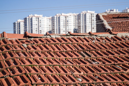 Weathered red tile roof of an old building contrasts with the backdrop of modern high rise residential buildings under a clear blue sky showing the intersection of urban transformation and history.の写真素材