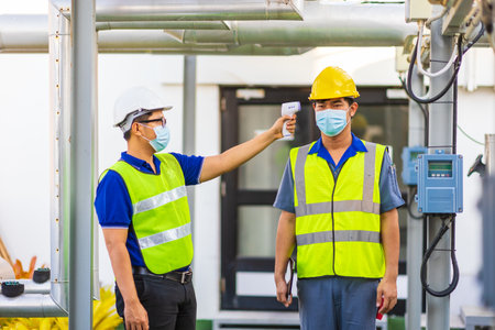 Asian male officer measuring temperature of engineer with thermometer at factory.の写真素材