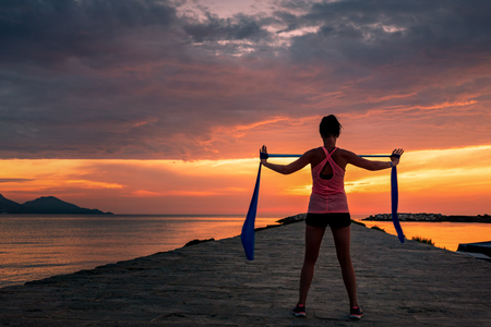Fitness woman celebrating fitness workout with rubber band - success and motivation towards the sea and sunset or sunrise.の写真素材