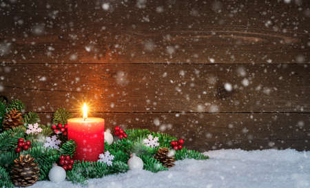 Christmas or Advent wood background with burning red candle on snow and snowflakes, decorated with fir branches and ornaments. Copy spaceの写真素材