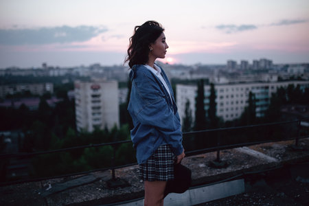 A girl in a hat and a denim jacket walks on the roof at sunset