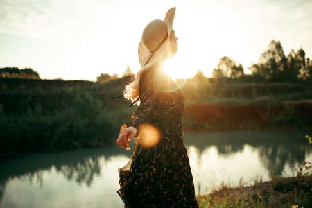 Stylish young woman in hat and dress standing near river at sunsetの写真素材