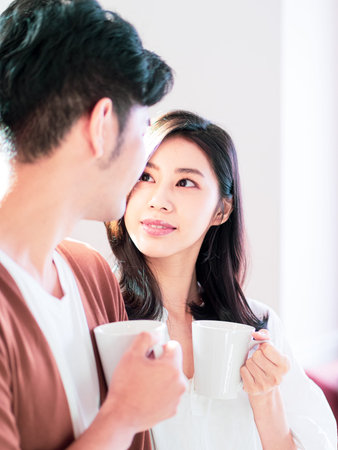 Young asian woman and man are enjoying spending time together at home with cup of coffee in hands.