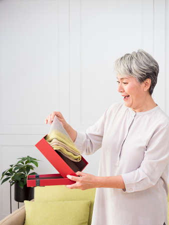 Portrait of an elderly Asian woman open gift boxes, smiling and looking down while standing in a living room at home.の写真素材