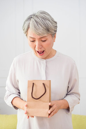 Portrait of an elderly Asian woman receives gift boxes, smiling and looking down while standing in a living room at home.の写真素材