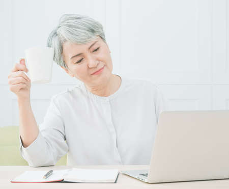 Senior asian woman using wireless laptop apps browsing internet and drinking coffee.の写真素材