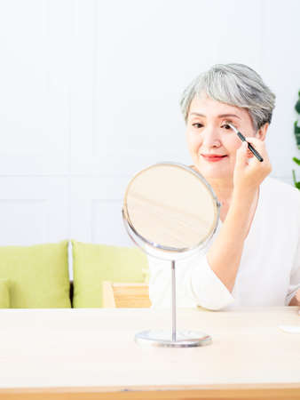 Senior asian woman applying foundation to her cheek with a makeup brush while sitting alone in front of a mirror.の写真素材