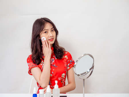 Portrait of a young asian woman in traditional cheongsam qipao dress looks at mirror and does skin care isolated on white background.の写真素材