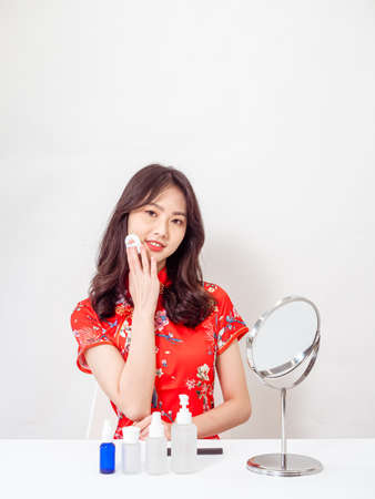 Portrait of a young asian woman in traditional cheongsam qipao dress looks at mirror and does skin care isolated on white background.の写真素材
