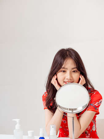 Portrait of a young asian woman in traditional cheongsam qipao dress looks at mirror and does skin care isolated on white background.の写真素材