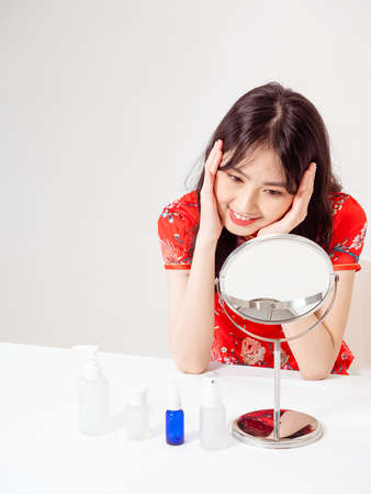 Portrait of a young asian woman in traditional cheongsam qipao dress looks at mirror and does skin care isolated on white background.の写真素材