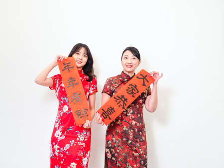 Asian women both wearing traditional cheongsam qipao dress hold Chinese festival couplet to celebrate new year with word meaning "May there be surplus year after year" on white background.の写真素材