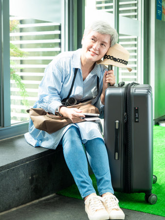 Portrait of senior adult elderly asian woman 60s smiling sitting with suitcase luggage bag and holding passport with ticket for travel concept.の写真素材