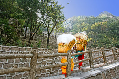 two women carrying heavy baskets up steep path by rock wall in China