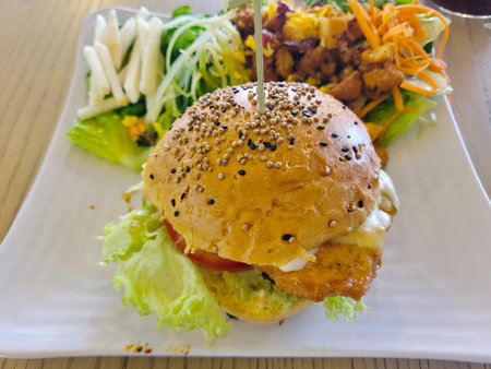 Savory chicken burger with a multigrain bun, fresh lettuce, and tomato, paired with a crisp side salad on a white plateの写真素材