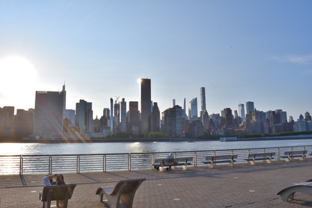 A New York city skyline with a river in the background from Roosevelt Island. People are sitting on benches and enjoying the viewの写真素材