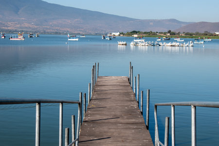 A wooden pier extends over the tranquil waters of Lake Cajititlan in Jalisco, Mexico, with boats and mountains in the backgroundの写真素材