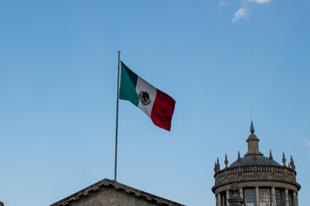 Guadalajara, Mexico - February 2 2025: Large Mexican flag waving near historic Cabanas building dome on a sunny day in Guadalajara, Jalisco, Mexicoの写真素材