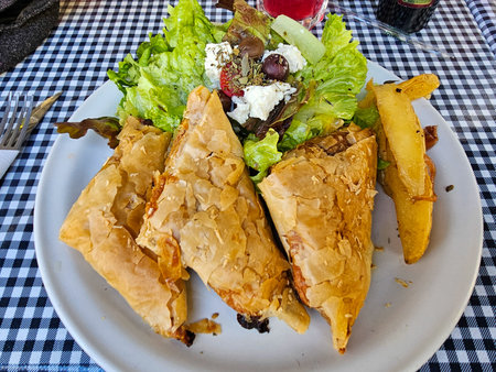 Close-up of a delightful Greek lunch with savory pastries, fresh salad (feta, olives), and crispy fries, on a checkered tablecloth.の写真素材