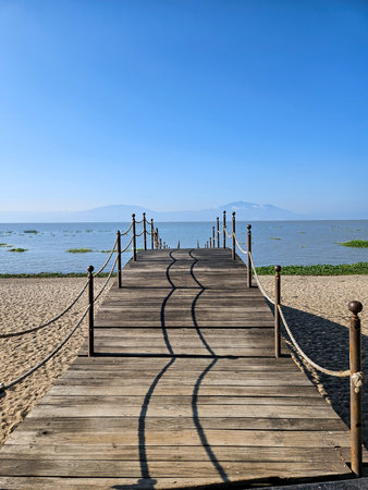Long wooden pier extending into a lake, on a bright sunny day. Features sand, water, and distant mountains under a clear blue skyの写真素材