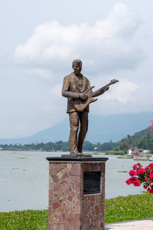 Chapala, Mexico - August 31 2025: Bronze statue of musician Micky Laure with electric guitar on a pedestal by Lake Chapala. Green vegetation and distant mountainsのeditorial素材