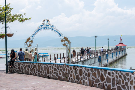 Chapala, Mexico - August 31 2025: Malecon de Chapala, Mexico, with people strolling the pier, an archway, a small lighthouse, and boats on the vast lake under a bright skyのeditorial素材