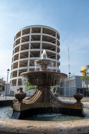 Guadalajara, Mexico - November 8 2025: A traditional stone fountain spraying water in the foreground, with a striking modern circular concrete building in the backgroundのeditorial素材
