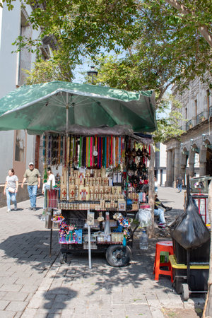 Guadalajara, Mexico - November 8 2025: Downtown Guadalajara street cart selling religious items, colorful necklaces, and souvenirs. Sunny day, green umbrella, historic architecture, pedestriansのeditorial素材