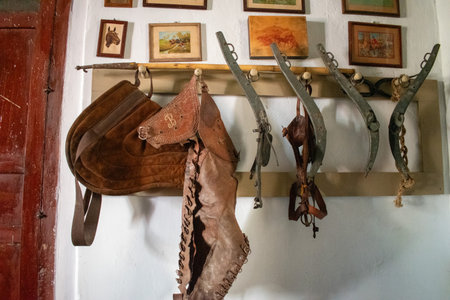 Cocula, Mexico - April 27 2025: Old leather saddles, stirrups and framed paintings displayed on the wall in Hacienda La Sauceda, Coculaのeditorial素材