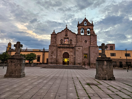 Zapopan, Mexico - July 13 2024: Historic San Pedro Apostol Parish Church in Zapopan, Jalisco. Stone facade, bell tower, crosses in plaza. Twilight under dramatic cloudy sky.のeditorial素材