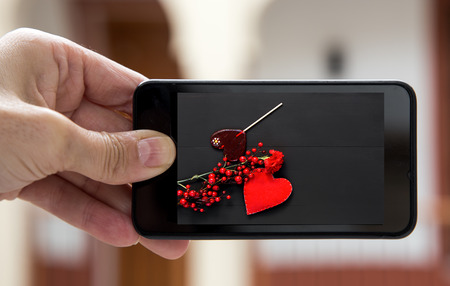 Hand holding smartphone with image of bouquet of red carnations and handmade red heart on black backgroundの写真素材