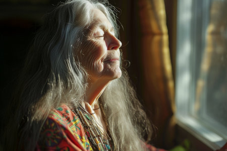 Generative AI illustration of older woman with long gray hair in her living room doing yoga and meditation with sunlight coming through a window. Relaxの素材