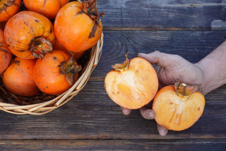 man holding persimmon fruit open in half next to a basket full of fruit with a wooden baseの写真素材