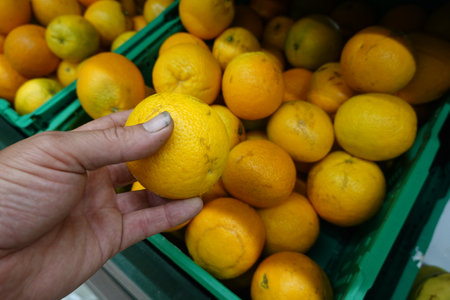 A close-up of a hand holding a fresh orange while selecting from a crate of oranges in a grocery store.の写真素材