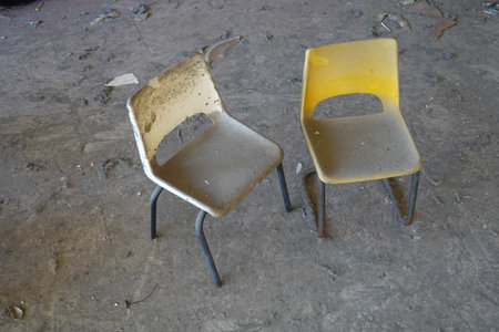 A pair of dusty, abandoned chairs in a neglected classroom with debris scattered on the floor.の写真素材