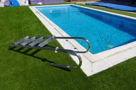 Grab bars and steps of a stainless steel pool ladder lying on artificial turf next to a home swimming poolの写真素材